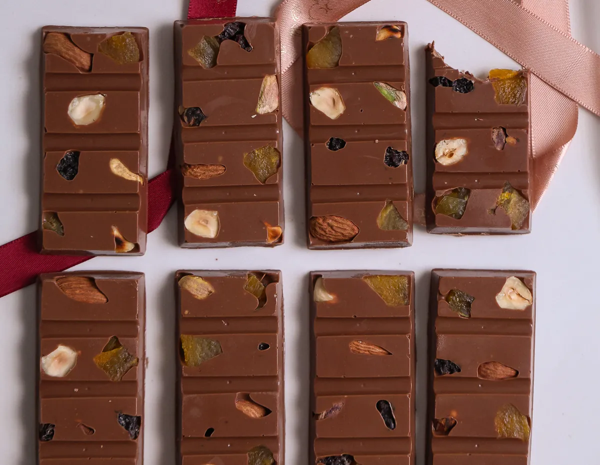 Barrinhas de chocolate ao leite com mix de nuts alinhadas na bancada, ao lado de fitas de cetim