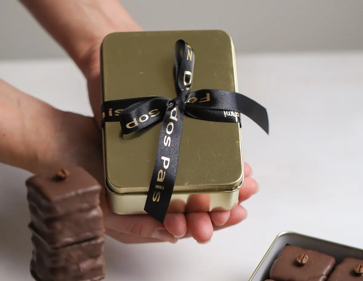 Mãos entregando uma lata dourada fechada com laço personalizado, com bombons sortidos ao lado